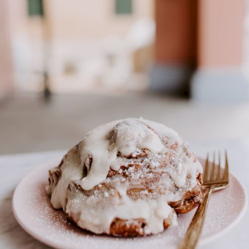 A cinnamon roll with icing on a pink plate next to a gold fork, set on a marble surface with a blurred background.