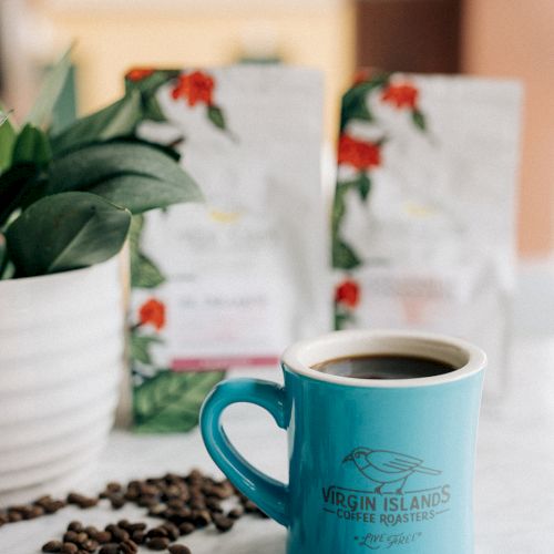 A blue mug of coffee, surrounded by coffee beans, sits on a table. Two coffee bags and a potted plant are in the background.