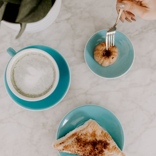 A hand holding a fork, a pastry, coffee in a blue cup, toast on a blue plate, and a plant on a marble surface.