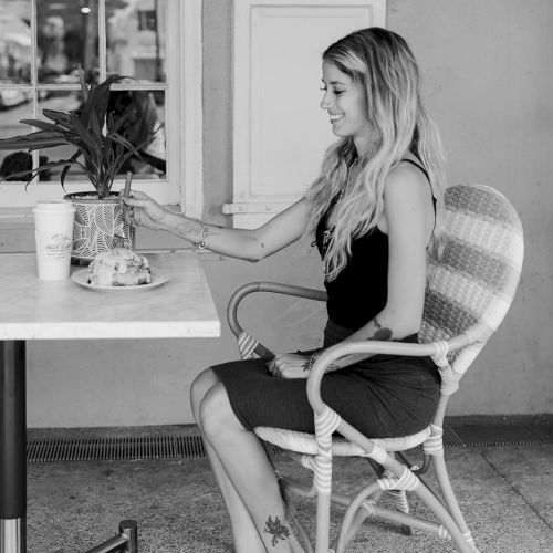 A woman sits at an outdoor table with a drink and a plant, wearing casual clothes and sneakers, in a black and white setting.