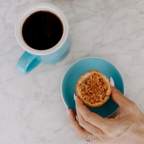 The image shows a hand holding a muffin over a blue saucer, next to a cup of coffee on a marble surface.