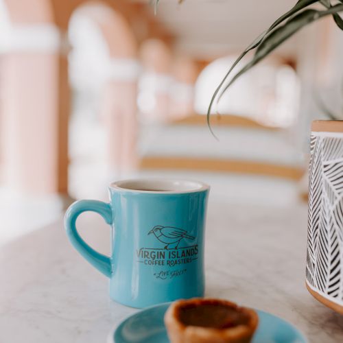 A blue mug and a small pastry on a blue plate sit on a marble table, with a plant and blurred background.