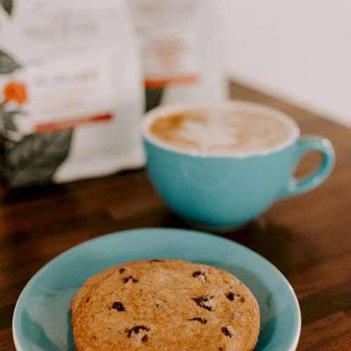 A chocolate chip cookie on a blue plate and a latte in a cup are on a wooden table with bags in the background.