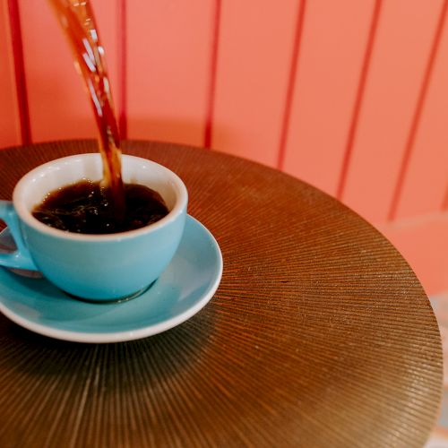 A stream of coffee is being poured into a blue cup and saucer on a textured table, against a red paneled background.