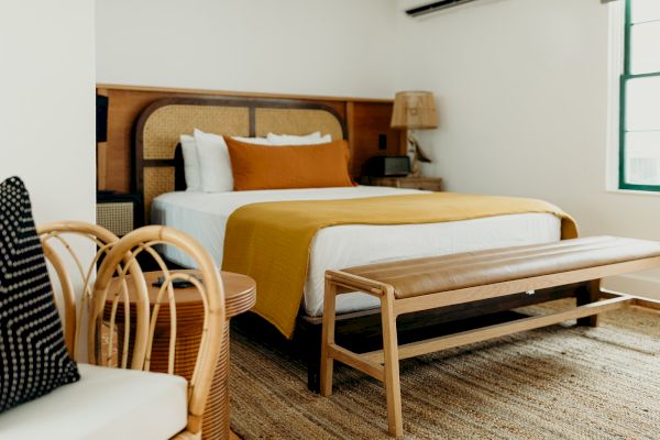 A cozy bedroom features a bed with mustard bedding, a woven headboard, a wooden bench, a rattan chair, and a window with green trim.