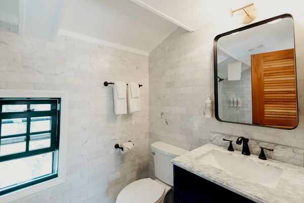 The image shows a bathroom with white marble tiles, a toilet, a dark vanity with a sink, a mirror, and a window with black trim.