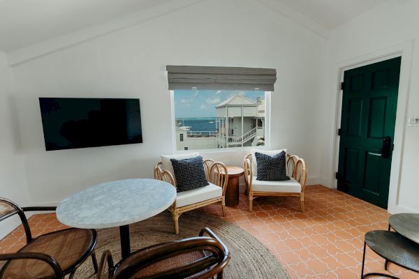 A cozy room with a mounted TV, round table, chairs, two cushioned seats by a window with an ocean view, and a dark green door.