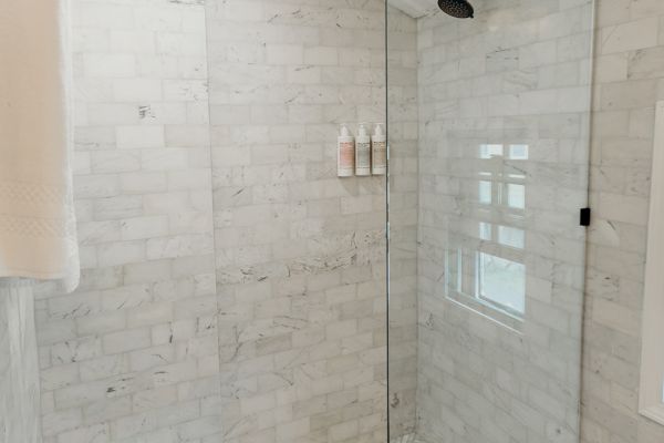 The image shows a modern shower with white marble tiles and a glass enclosure. Black showerhead and patterned floor tiles complete the look.