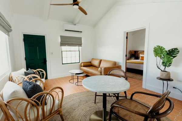 A cozy, modern room with wicker chairs, a sofa, round tables, and a plant; leads to a bedroom with white walls and terracotta floors.