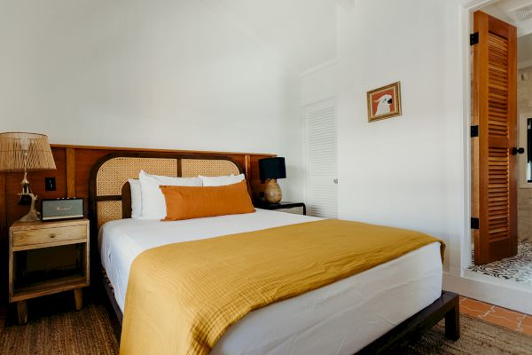 A cozy bedroom with a bed, white and mustard bedding, bedside tables, lamps, and a partially open door to another room.