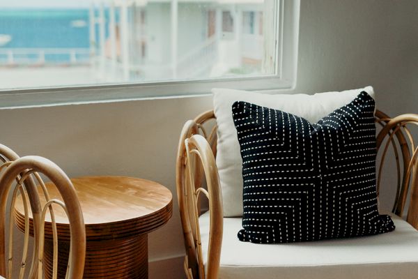 The image shows a cozy sitting area with wicker chairs, a patterned cushion, a small wooden table, and a seaside view through a window.