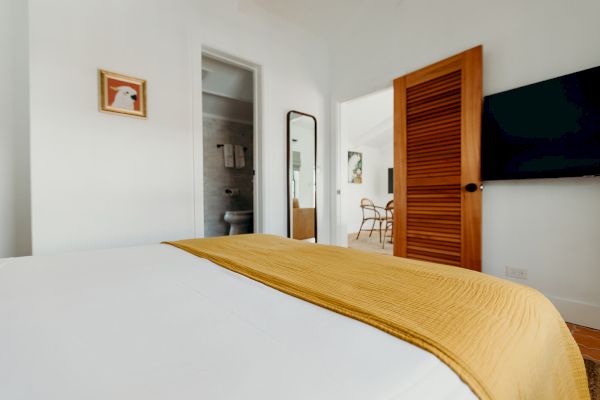 A bedroom with a bed, yellow blanket, a TV, and a wooden door; artwork and a mirror are on the walls, with a view into the bathroom.