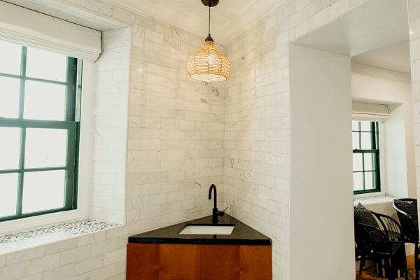 A small corner sink with a wooden cabinet, pendant light, and patterned tile floor in a room with brick walls and a window.