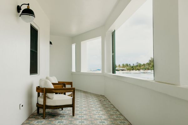 A balcony with two wooden chairs, white walls, patterned tile flooring, overlooking a scenic view of the ocean and trees in the distance.