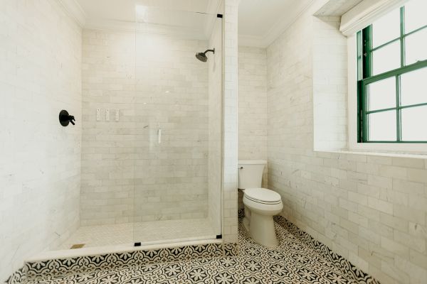 The image shows a bathroom with a glass-enclosed shower, patterned tile floor, white brick walls, a toilet, and a window with green trim.