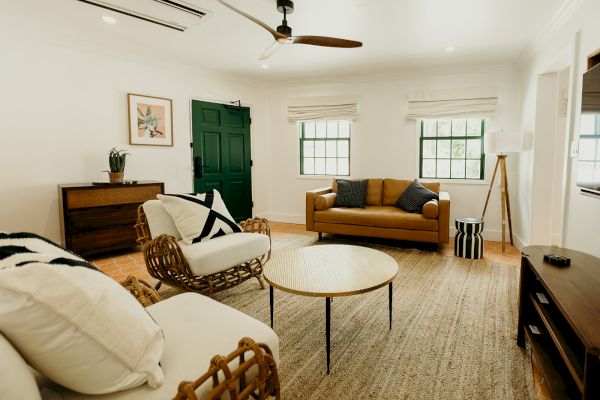 A cozy living room with modern decor, featuring a brown sofa, wicker chairs, a round table, a green door, and natural light.