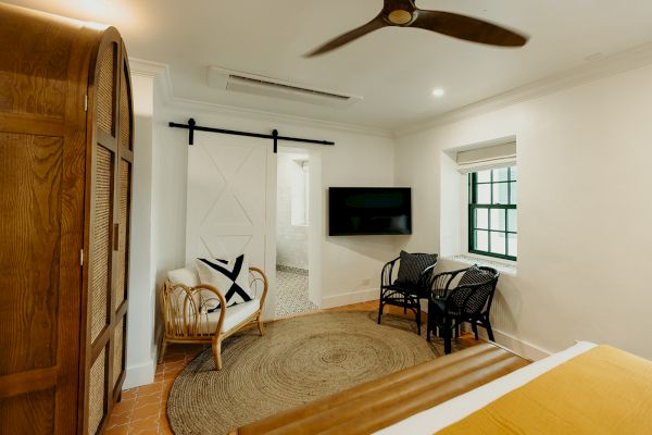 A cozy room with a bed, wicker chairs, a wooden wardrobe, TV, ceiling fan, and a sliding door leading to a bathroom.