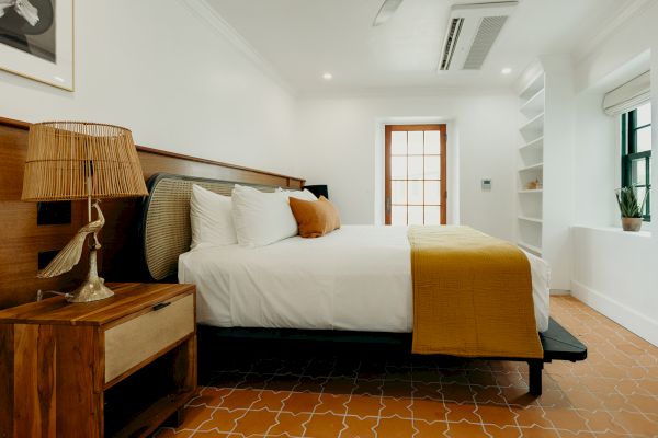 A cozy bedroom with a large bed, white and mustard bedding, a wooden nightstand with a lamp, open shelves, and a small plant by the window.