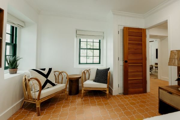 A cozy room with two wicker chairs, cushions, a small table, plant, and terracotta tiled floor. Doorway leads to another room.