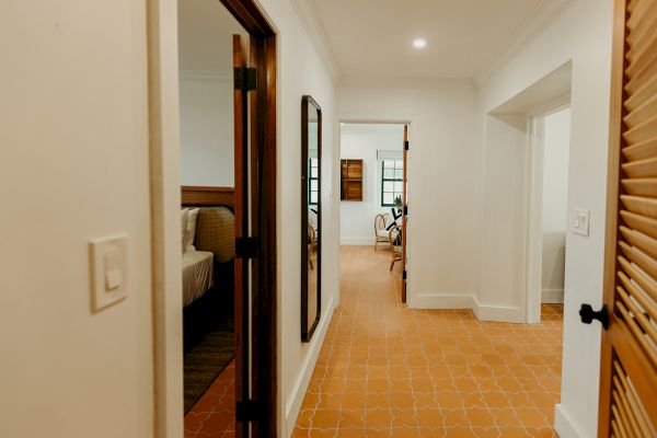 A hallway with tile flooring, leading to a bedroom and living area with wooden furniture and white walls, captured in warm lighting.