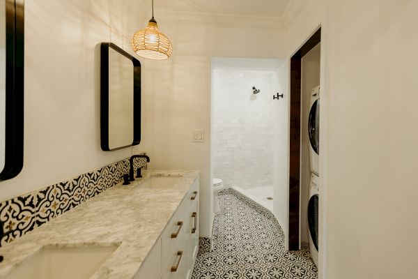 A bathroom with a marble countertop, patterned tiles, a shower, and a stacked washer and dryer combo unit in the corner.