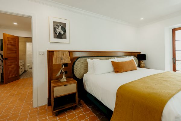 A cozy bedroom with a large bed, orange blanket, wooden headboard, beside lamps, and open door to a bathroom, in a warmly lit space.