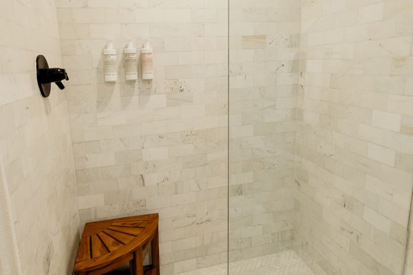 A modern shower with white tiles, a glass door, black fixtures, a wooden corner stool, and a few bottles on a wall rack.