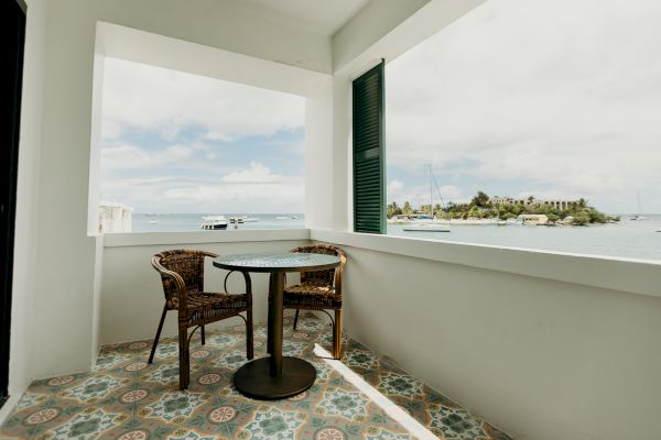 A small balcony with two wicker chairs and a round table overlooking the sea. There’s a tiled floor and a view of distant boats and an island.