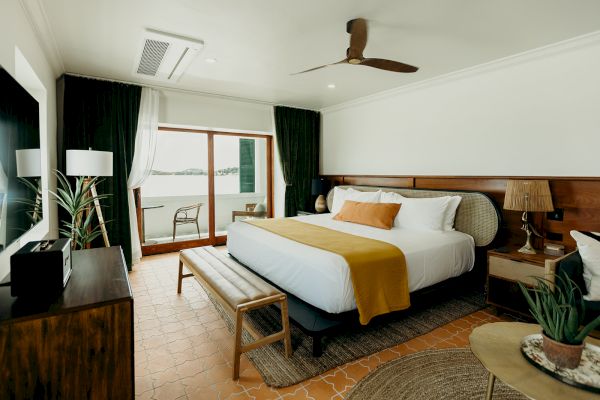 A cozy bedroom with a large bed, ceiling fan, plants, and a view through the sliding glass door, creating a warm and inviting atmosphere.