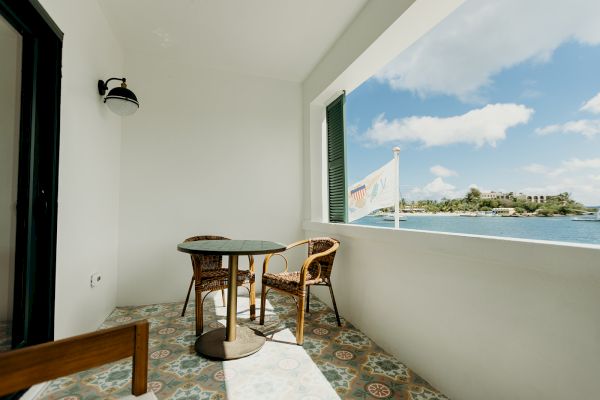 A small balcony with two chairs and a table, overlooking a body of water and distant buildings, with a pleasant, sunny sky.