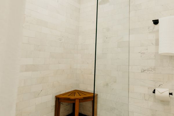 A modern shower with white tiles, a glass door, a large rainfall showerhead, and a small wooden bench in the corner.