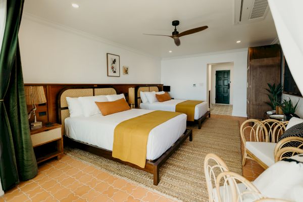 The image shows a stylish hotel room with two double beds, modern decor, a ceiling fan, and rattan furniture, creating a cozy atmosphere.