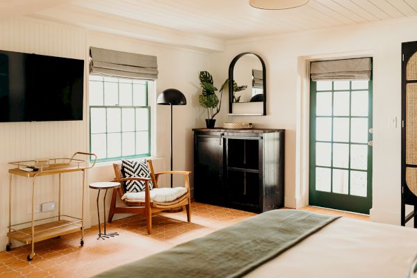 A cozy room with a large window, armchair, TV, and a cart. Features a dark cabinet with a mirror and plants, and a door with glass panels.