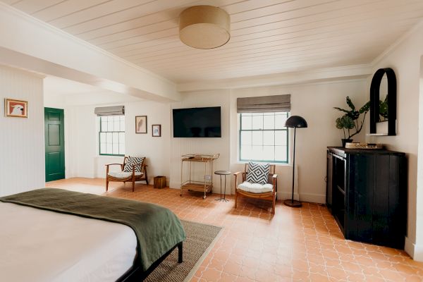 A cozy bedroom with a bed, TV, armchairs, and decor on light-colored walls and floor.