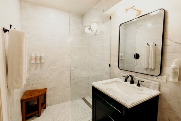 A modern bathroom with a glass shower, marble sink, large mirror, and towels. There’s a wooden bench and toiletries in the shower area.