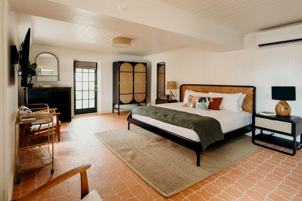 A cozy bedroom with a large bed, TV, chairs, wardrobe, and a rug on tiled flooring. The room features natural tones and a door to outside.