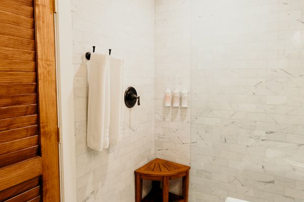 A bathroom featuring a wooden door, white tiled walls, a towel on a rack, a wooden stool, and decorative patterned floor tiles.