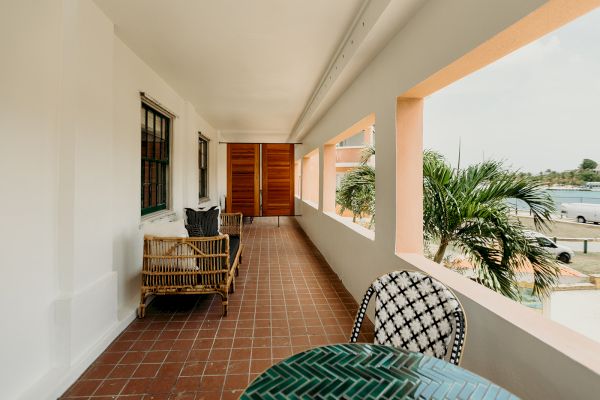 A veranda with wicker furniture, a table, and chairs overlooks a palm tree and waterfront view, with white walls and tile flooring.