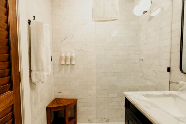 A small bathroom featuring a walk-in shower, wooden bench, marble counter, wall-mounted lights, and decorative floor tiles.