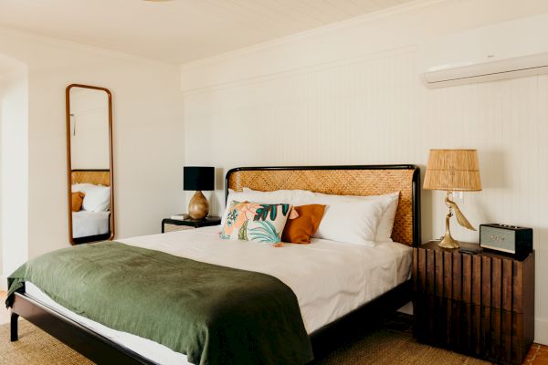 A cozy bedroom with a double bed, green blanket, decorative pillows, and wooden side tables. A mirror and air conditioner are on the wall.