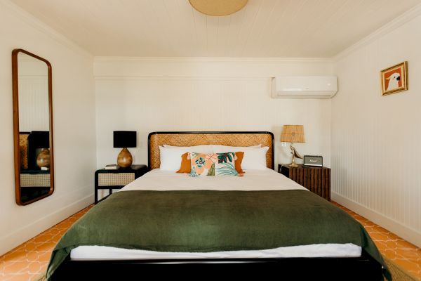 A bedroom with a neatly made bed, green blanket, decorative pillows, bedside tables, lamps, wall art, mirror, and an air conditioner on white walls.