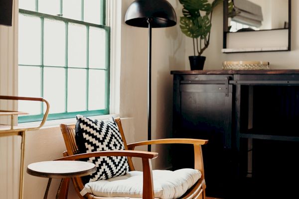 A cozy room with a wooden chair, geometric pillow, small table, black cabinet, tall lamp, plant, and window with Roman shade, under soft lighting.