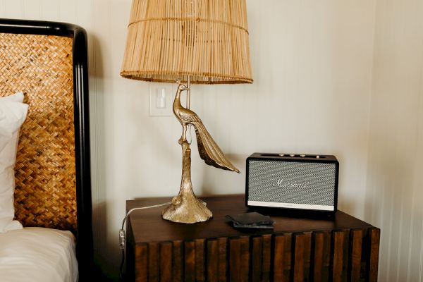 A decorative lamp with a bird base and a speaker are on a wooden bedside table next to a woven headboard.