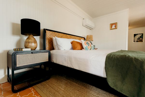 A cozy bedroom featuring a bed with pillows, a bedside table with a lamp and books, and a green blanket on a woven rug.