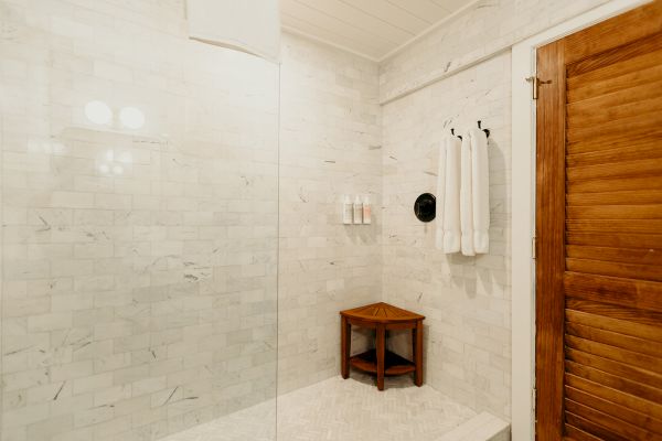 The image shows a shower with white tiled walls, a wooden bench, towels, and a glass divider, adjacent to a wooden door.