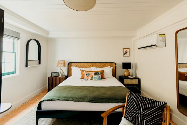 A cozy bedroom with a bed, stylish lamps, a chair, and a wall mirror. The room has warm lighting and a modern, minimalist design.