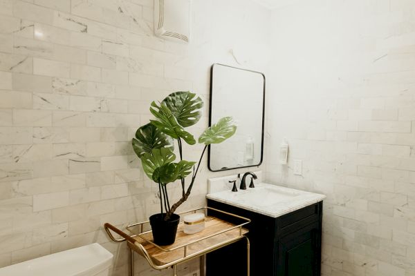 A small bathroom features a toilet, a sink with a mirror, a decorative plant on a wooden cart, and black and white patterned floor tiles.