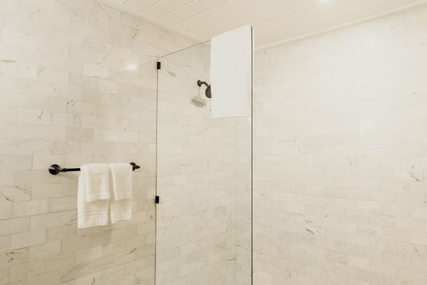 The image shows a modern shower area with glass walls, white tiles, and patterned floor tiles, featuring a towel hanging on a black rod.