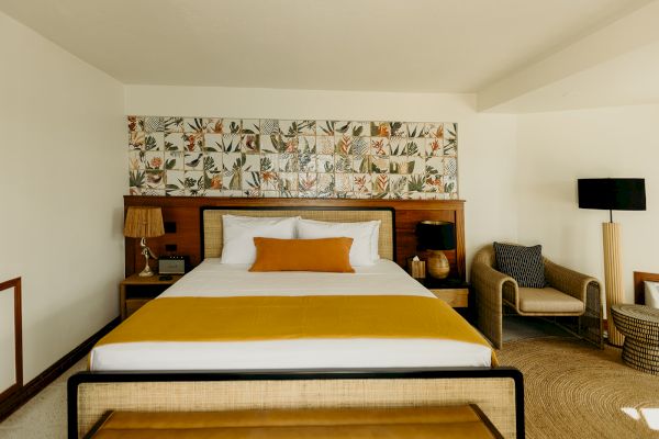 The image shows a bedroom with a large bed, colorful headboard, lamps, a chair, and a rug, in a cozy, well-decorated space.